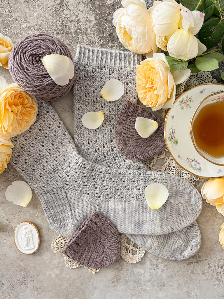 A top-down photo of a pair of light blue-gray socks crossed over each other at the toe. They use the round toe construction, and two more light purple round toes are sitting nearby along with a purple yarn cake. Everything is surrounded by a bunch of yellow roses, and a teacup full of tea is off to the right side.