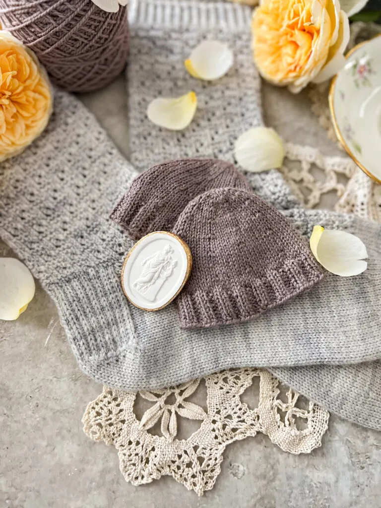 A flatlay photo showing two light purple swatches laid on top of a pair of light blue-gray handknit socks. The purple swatches show round toes knit in two different ways. Leaning against them is a small plater intaglio.