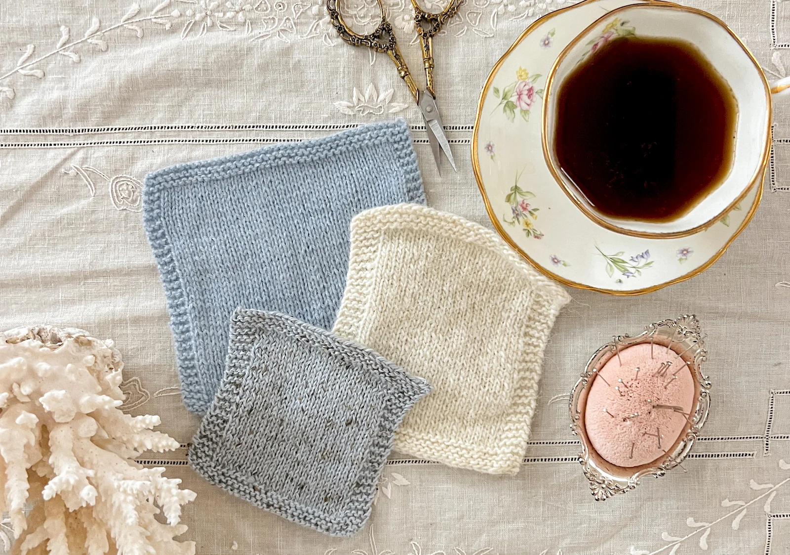 A flatlay photograph showing three swatches of yarn in blue, white, and gray, along with a chunk of white coral, a pair of brass scissors, a teacup full of coffee, and a pink pincushion.
