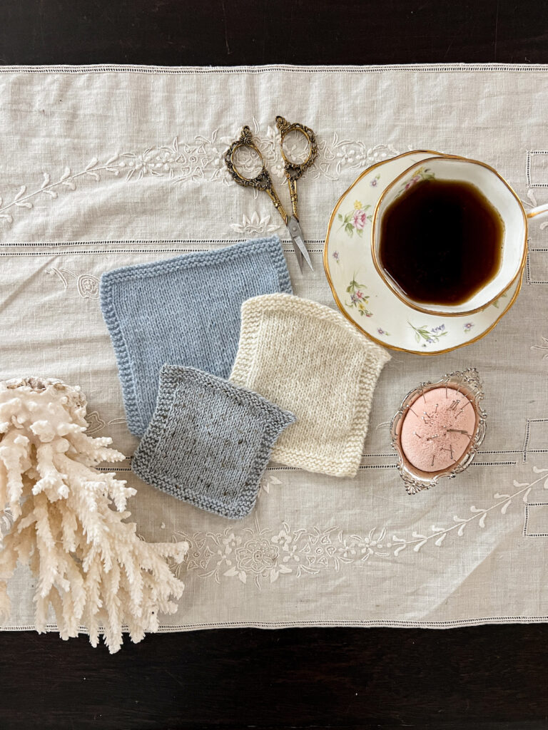 A flatlay photograph showing three swatches of yarn in blue, white, and gray, along with a chunk of white coral, a pair of brass scissors, a teacup full of coffee, and a pink pincushion.