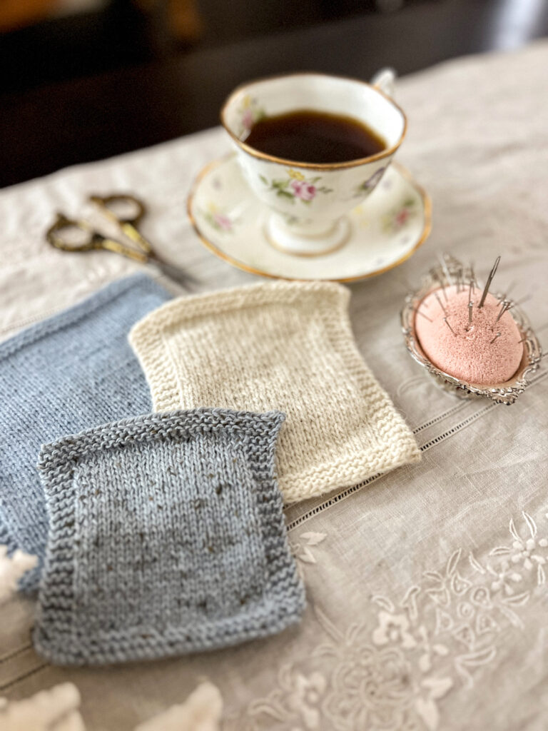 A low-angle photo with a swatch of gray yarn in the foreground, knit with a woolen spun yarn. Blurred in the background are a blue swatch and a white swatch, along with some brass scissors, a white teacup full of coffee, and a pink pincushion.