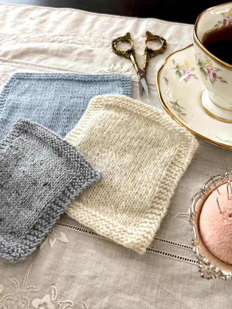 A photo showing three swatches of yarn. In the foreround are a white swatch knit with a single-ply yarn and a gray swatch knit with a woolen-spun yarn. Blurred in the background is a blue swatch knit with a worsted-spun yarn.