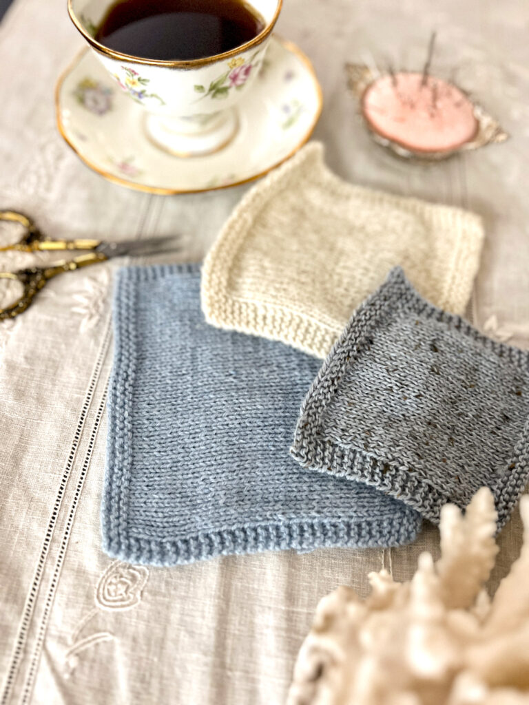 Three yarn swatches are laid flat on an antique table cloth. Only the blue swatch, knit in a worsted-spun yarn, is in focus. Blurred in the background is a pastel teacup full of espresso and a pink pincushion.