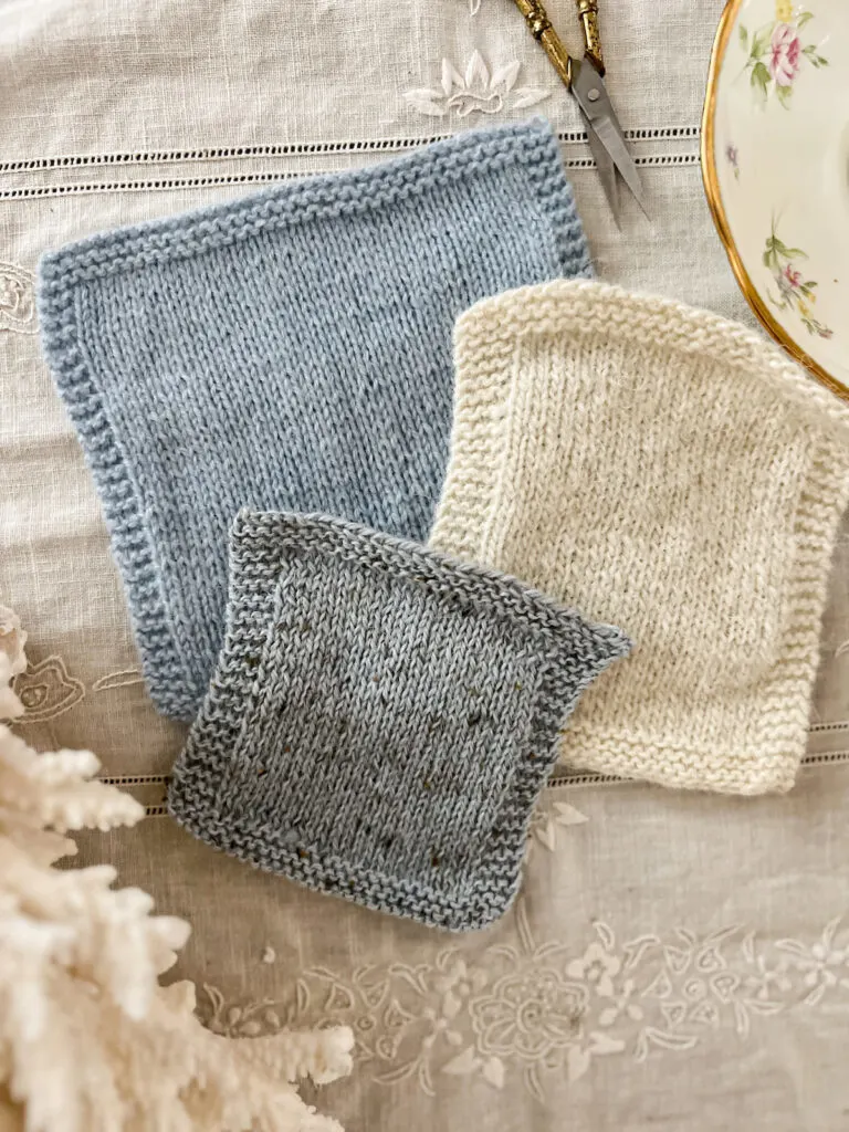 Three swatches knit in three different yarn constructions are laid flat on an antique tablecloth. The top swatch, in blue, is a worsted-spun yarn. The middle swatch, in white, is a single-ply yarn. The bottom swatch, in gray, is a woolen spun yarn.
