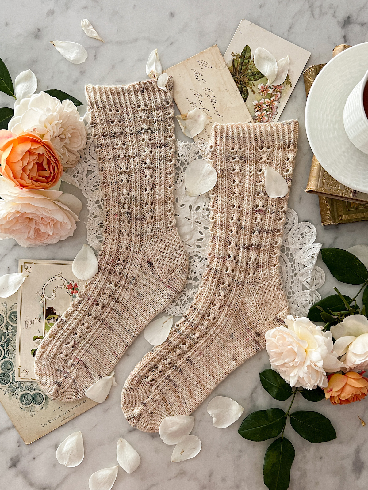 A pair of lightly speckled, pale pink socks is laid flat on a white marble countertop. The socks have columns of eyelets and purl stitches. They're surrounded by roses and antique paper ephemera.