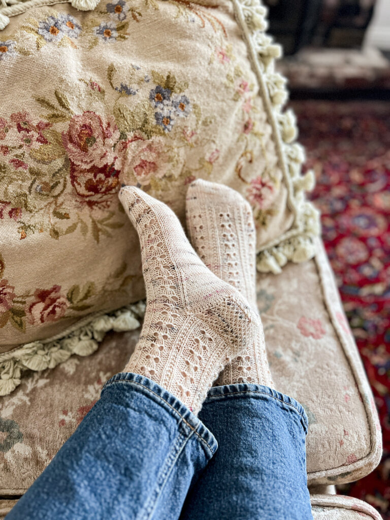 A pair of lightly speckled, pale pink socks on two feet lightly crossed at the ankle. The left foot, resting on top, is angled so the heel flap on the sock is visible.