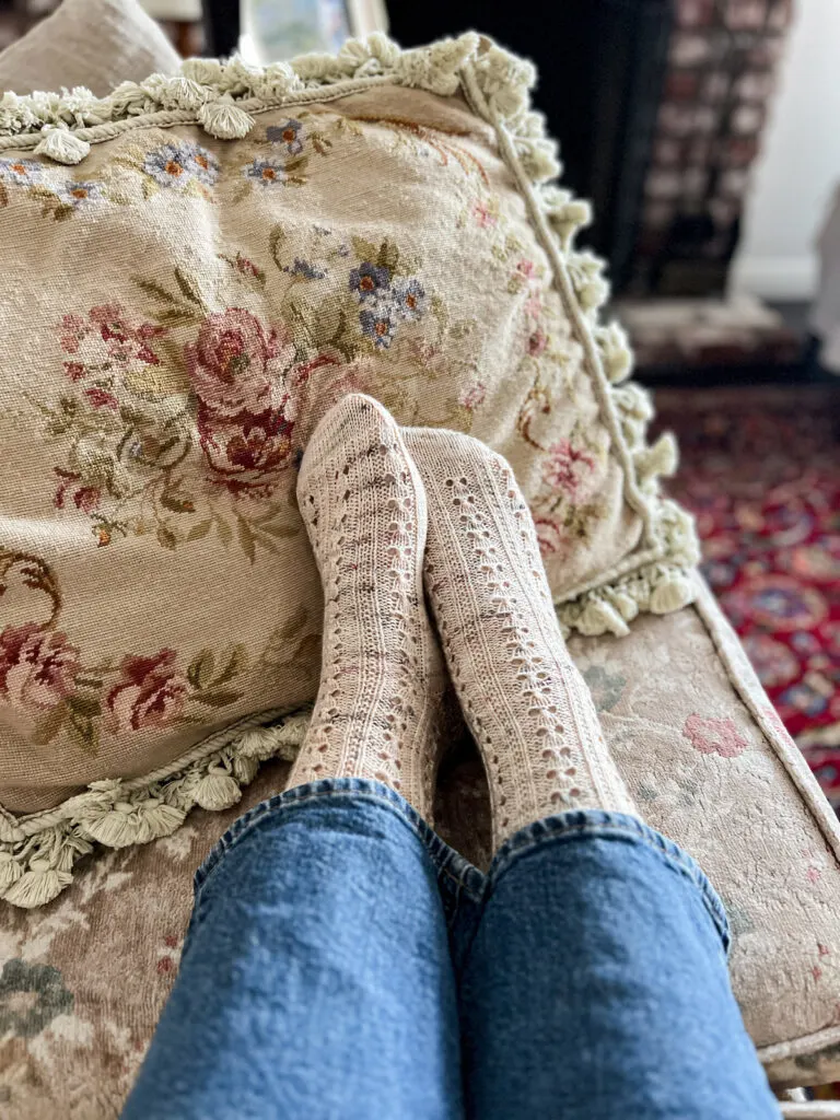 A pair of lightly speckled, pale pink socks on two feet resting against each other in front of an Aubusson-style pillow.