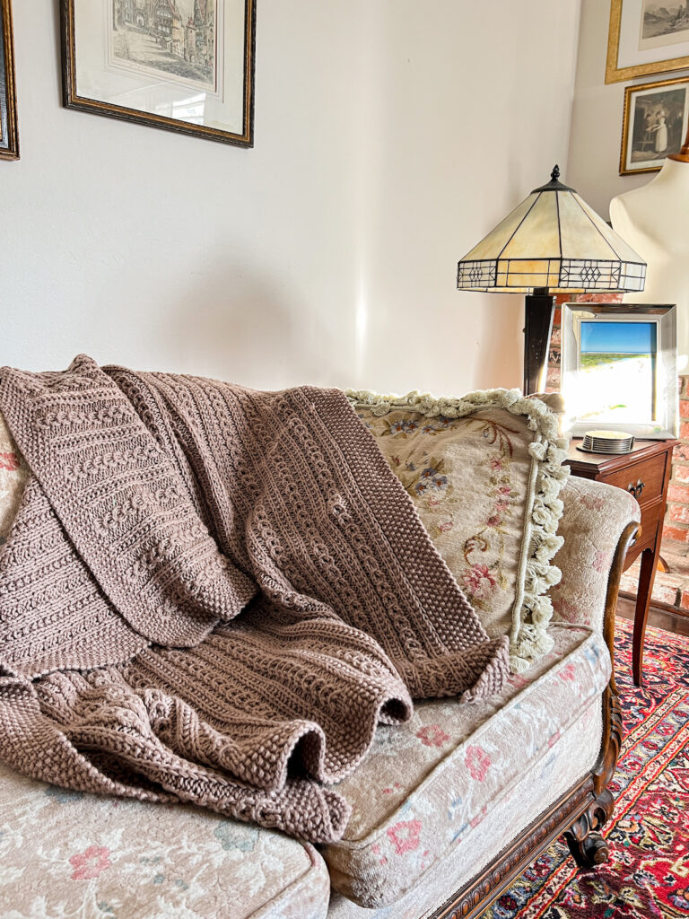 A brown blanket with columns of faux cables is draped across an antique sofa with floral upholstery.