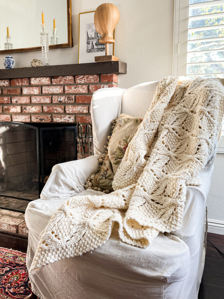 A cream-colored knit blanket is draped across a wingback chair with a white slipcover on it. The blanket has lots of seed stitch and interlocking lacy panels.