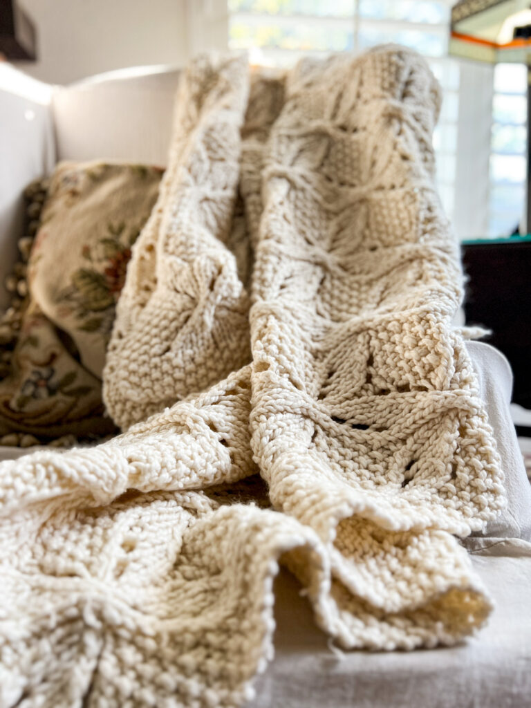 A photo of a handknit blanket made with chunky acrylic yarn that has been draped across a white chair. The foreground and background are blurred. The middle is in focus and shows how well the yarn has held up over more than a decade of use.
