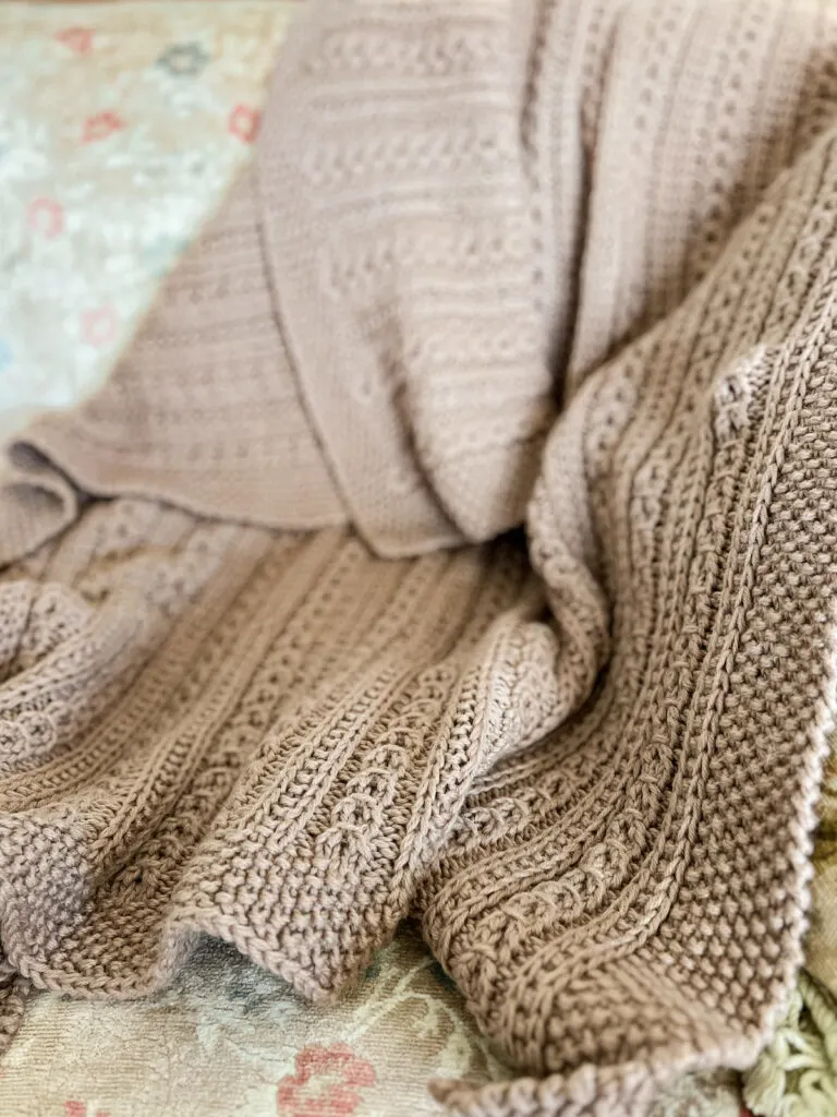 A brown handknit blanket with faux cables is draped across an antique sofa with floral upholstery. Only part of the blanket, in the foreground, is in focus. This shows how well the yarn has held up.