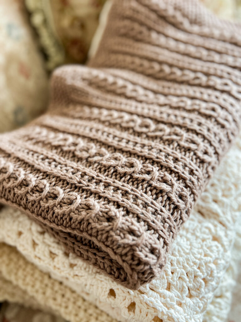 A pile of handknit and crocheted blankets. The top blanket, in brown with faux cables, is in focus while the others are blurred.
