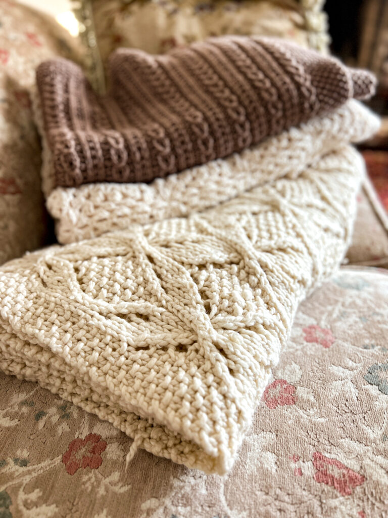 A pile of handknit blankets in cream and brown. The bottom blanket, knit from a chunky acrylic, is in focus in the foreground while the others are blurred.