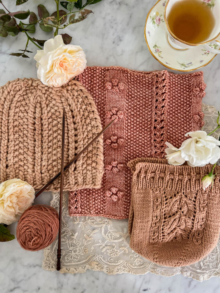 A pink knit hat, a pink knit cowl, and a pink knit tea cozy are all laid flat on a white marble countertop, surrounded by knitting needles, a teacup full of green tea, and some roses.