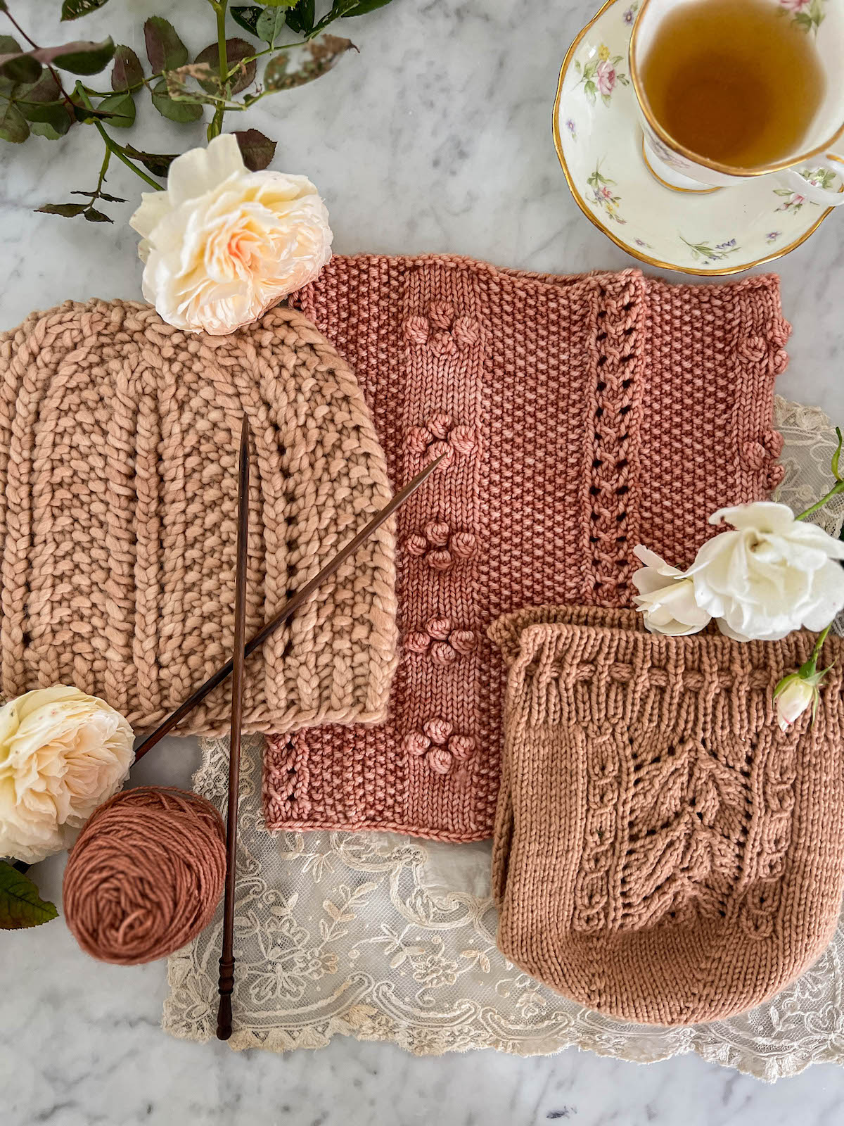 A pink knit hat, a pink knit cowl, and a pink knit tea cozy are all laid flat on a white marble countertop, surrounded by knitting needles, a teacup full of green tea, and some roses.