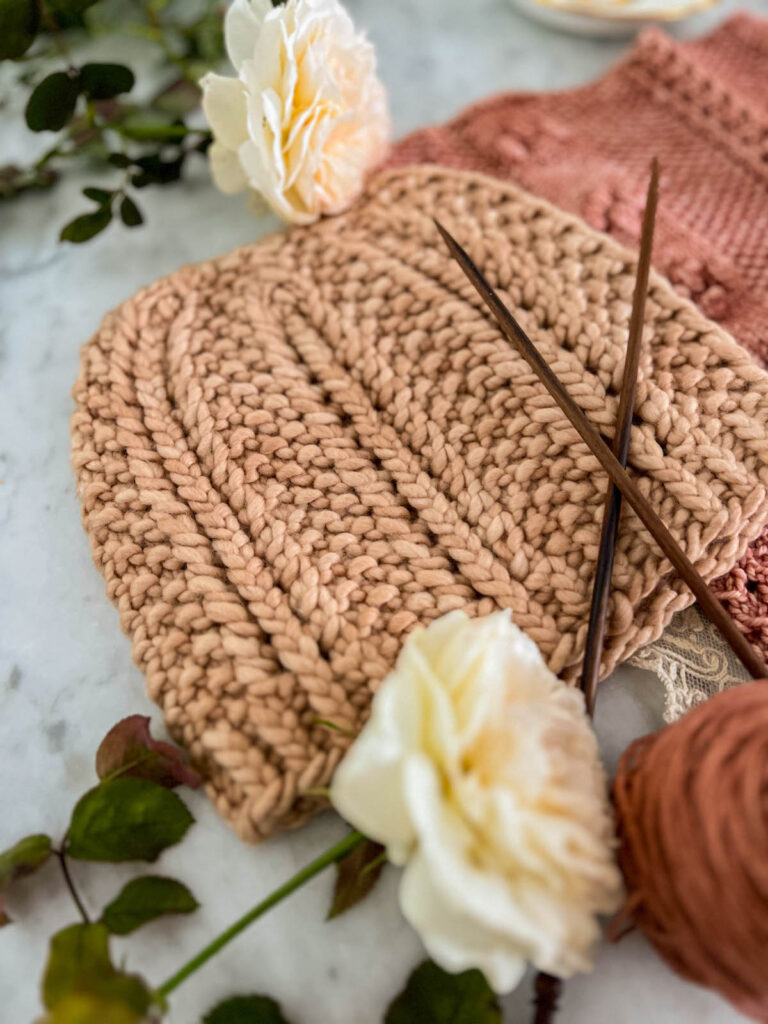 A pink hat with columns of eyelets and seed stitch is laid flat on a white marble countertop and photographed from a low angle.