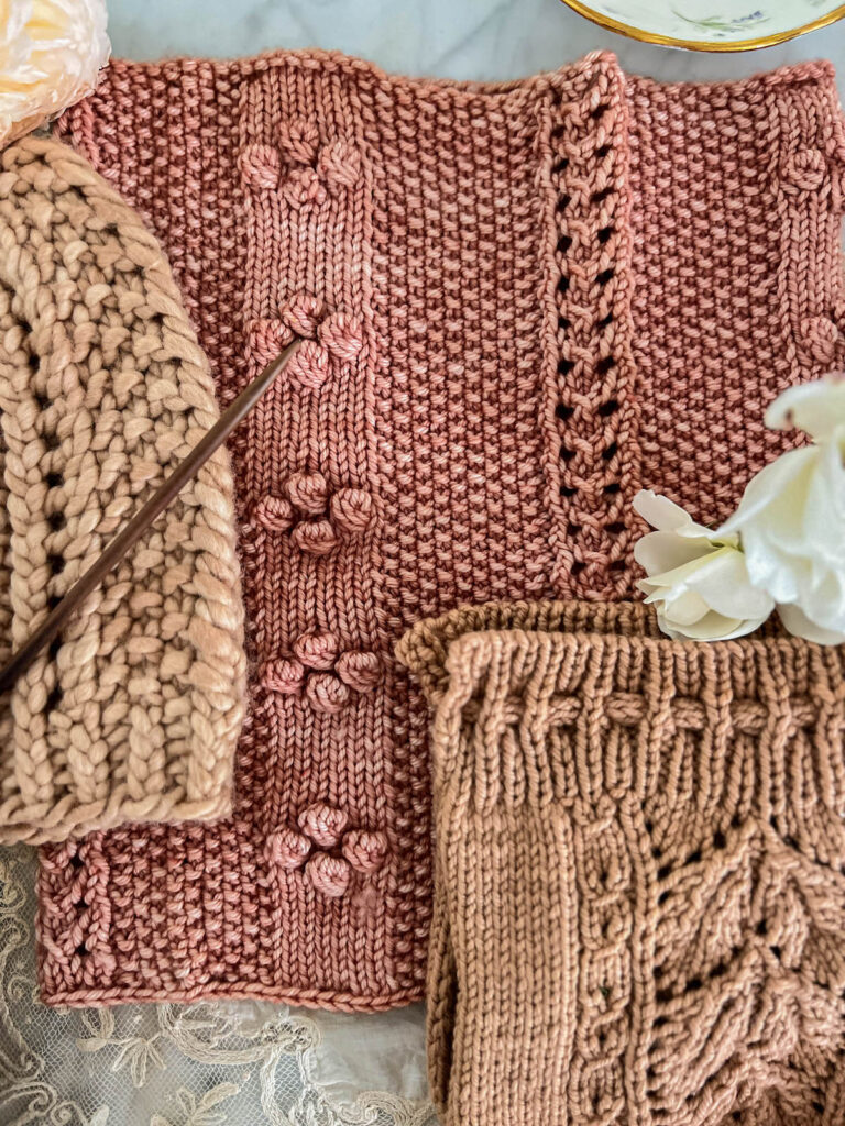 A pink knit cowl with columns of eyelets, bobbles, and seed stitch is laid flat on a white marble countertop. The edges of a pink knit hat and a pink knit tea cozy are visible to either side.