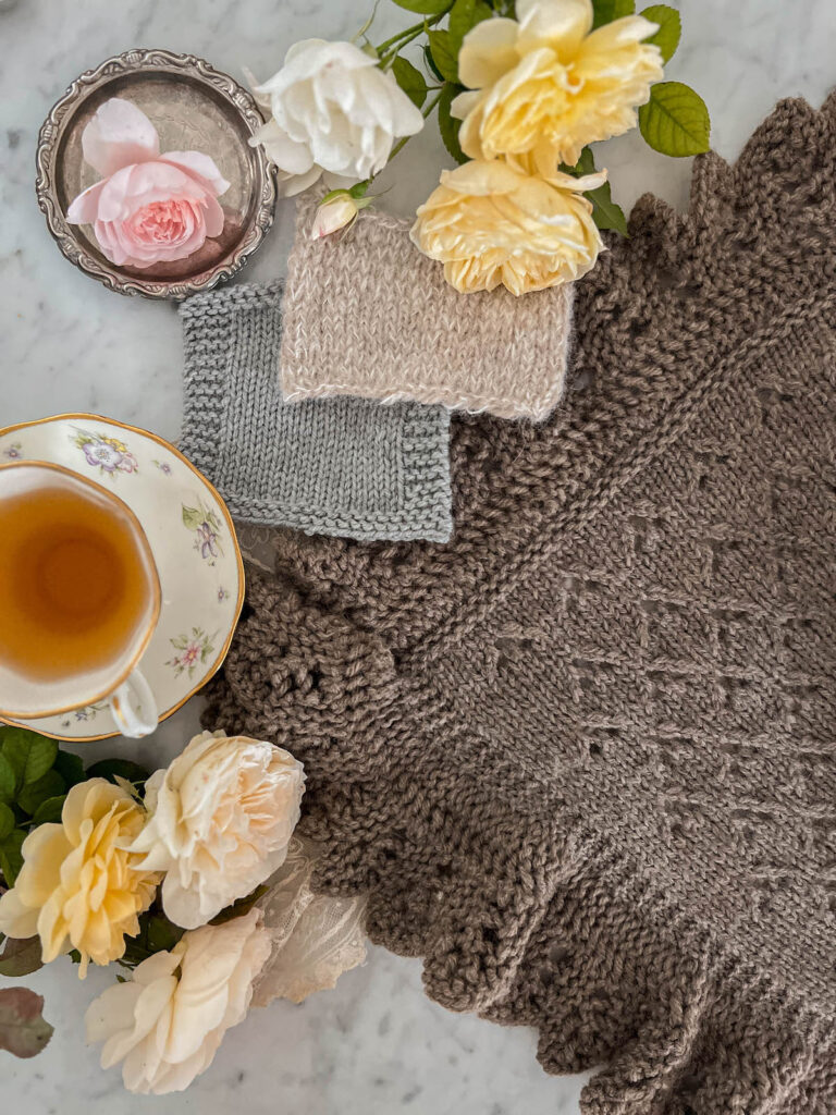 A top-down image showing two swatches of stockinette knitting on top of a large stockinette banket with an elaborate lace edging. To the left are a teacup full of tea and some roses.