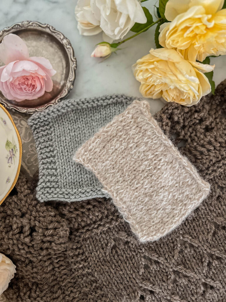 A close-up photo showing two knit swatches in stockinette stitch. One has a garter stitch border and one does not. They are on top of a larger piece of stockinette knitting. At the top of the image are some yellow, white, and pink roses.