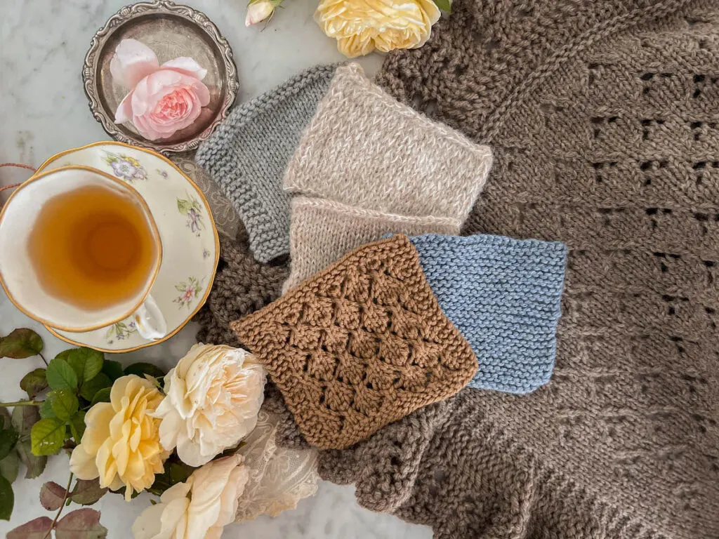 A top-down photo showing several swatches of knit fabric in garter stitch, stockinette, and simple lace to demonstrate how different techniques prevent the edges from curling. They are sitting on top of a larger piece of stockinette knitting. To the left are a white teacup full of tea and several yellow, white, and pink roses.