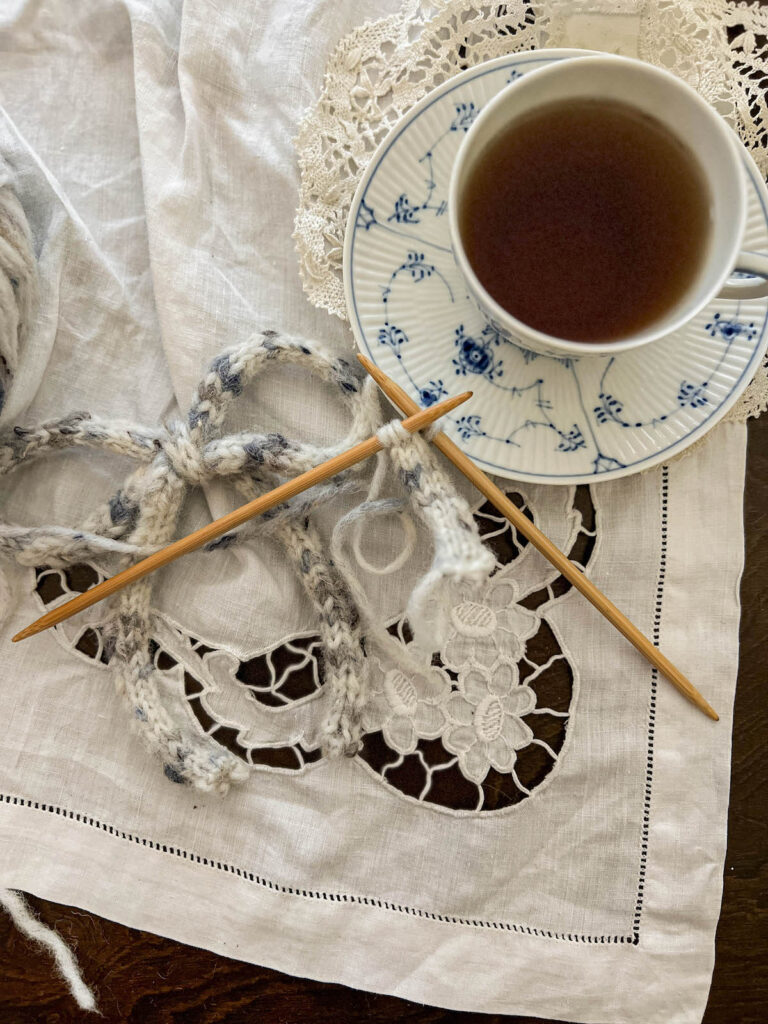 A flatlay photograph showing two wooden knitting needles with an i-cord in progress on them, an icord bow underneath, and a blue and white teacup full of tea in the top right corner.