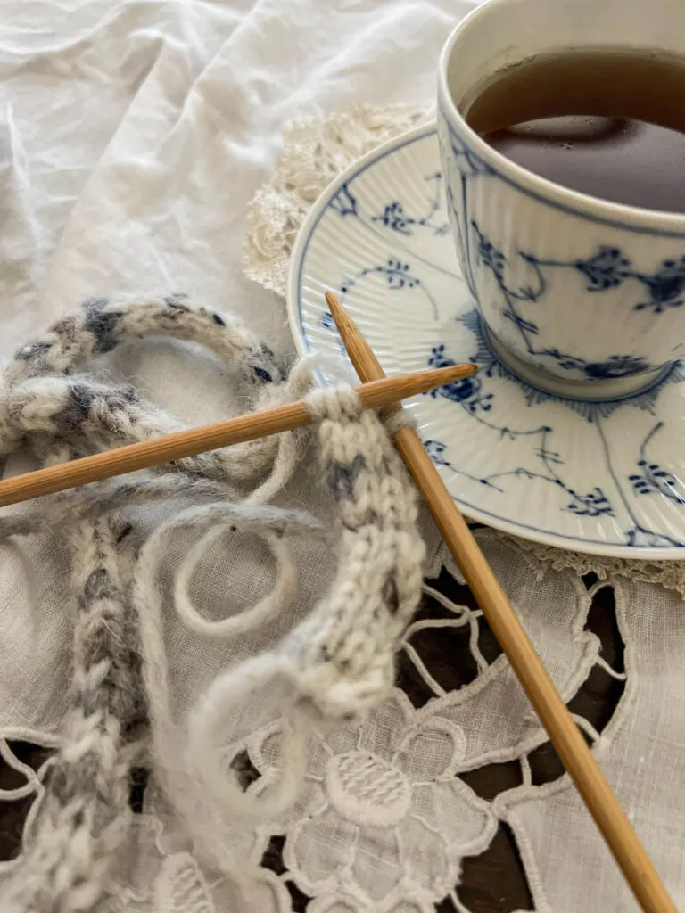 A close-up image showing the tips of two wooden needles with an i-cord in progress on them. They rest on the saucer of a blue and white teacup full of tea.