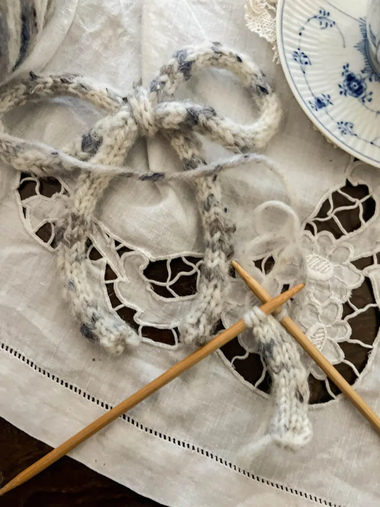 A flatlay image showing, in the top left corner, a bow tied from an i-cord. In the bottom right corner are two wooden knitting needles with another i-cord in progress on them.