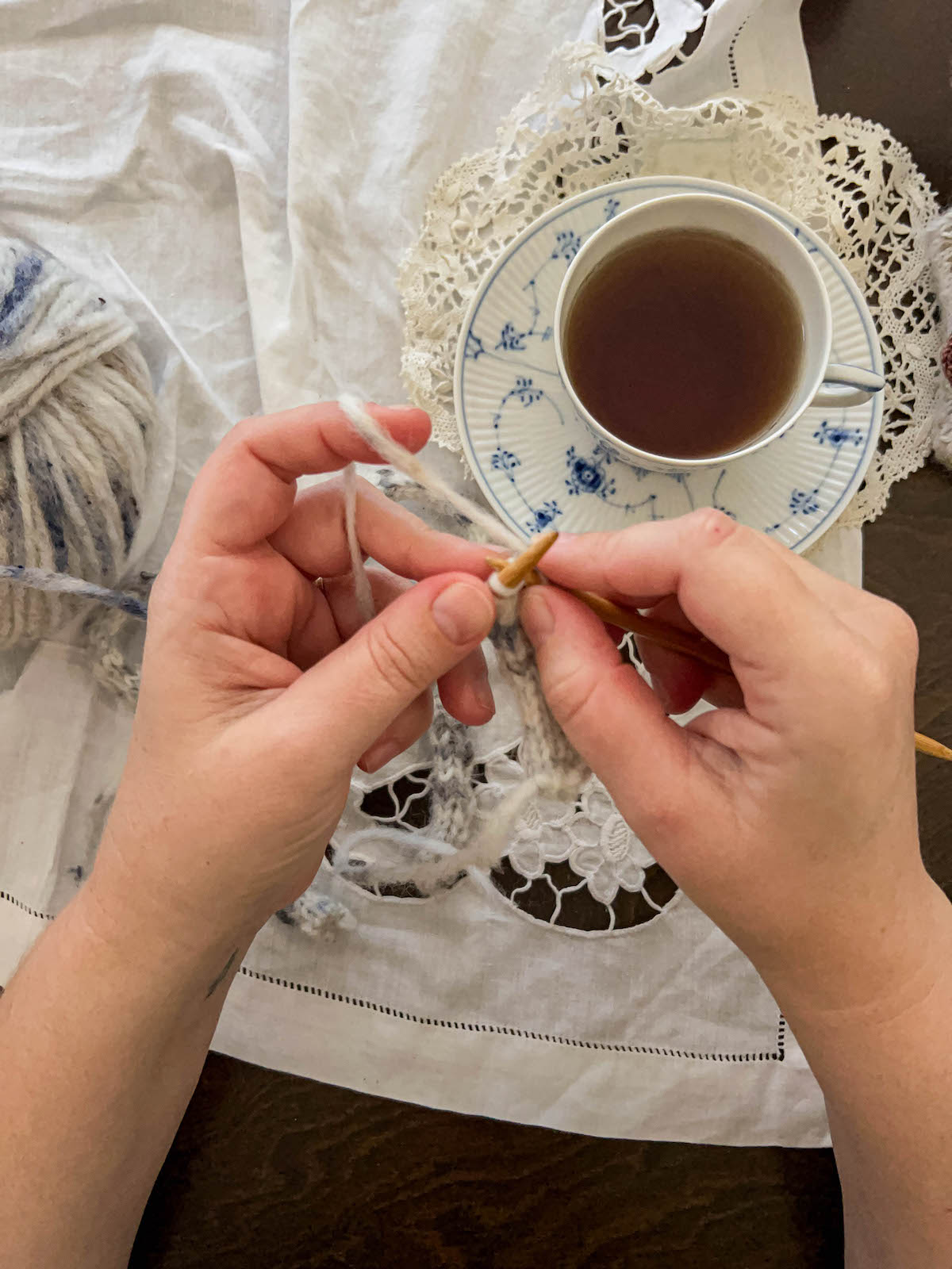 A white woman's hands (mine!) hold two wooden knitting needles while they work on making an i-cord. In the background on the table is a ball of yarn and a teacup full of tea.