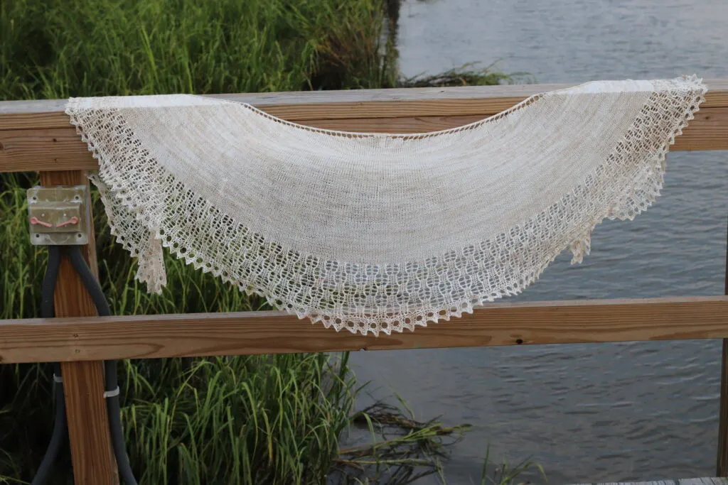A creamy crescent-shaped shawl with a stockinette body and wide lace edging is draped along a wooden fence rail.