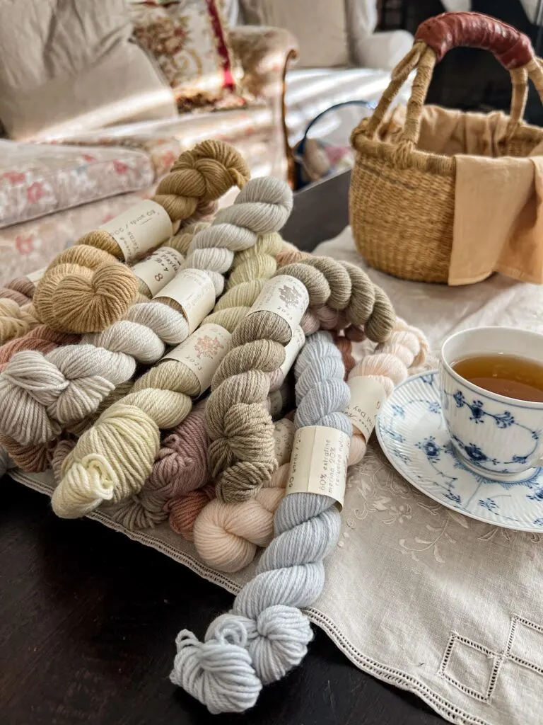 A side angle photo of a pile of pastel mini skeins of yarn. A blue and white teacup sits to the right of the pile while a grass basket sits empty in the background.