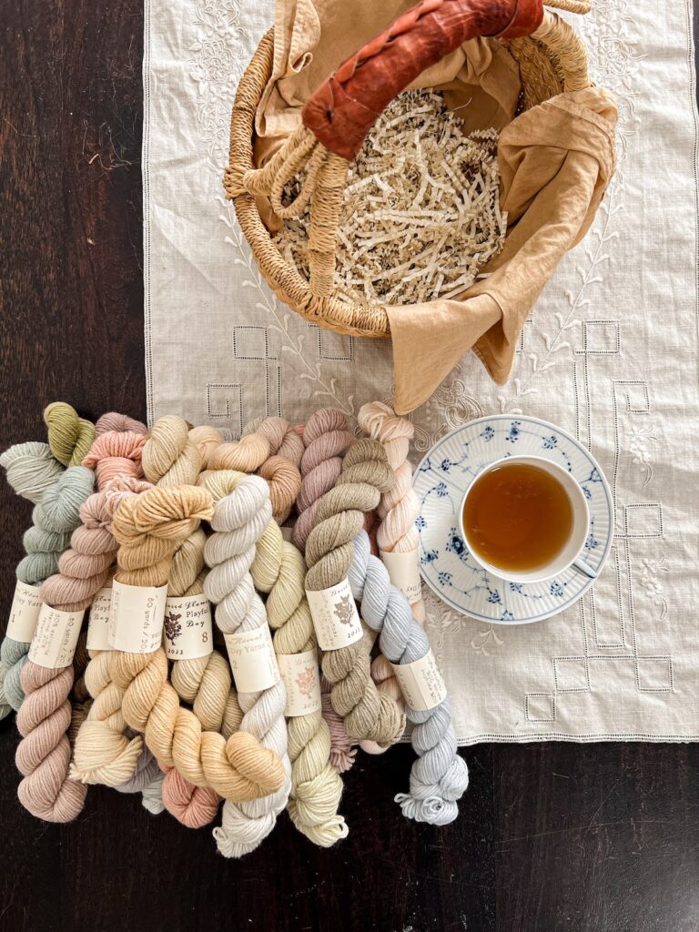 A top-down photo showing a pile of pastel mini skeins of yarn sitting on a dark brown coffee table. Next to the pile is a blue and white teacup full of coffee and above it is a grass basket full of crinkled paper shavings.