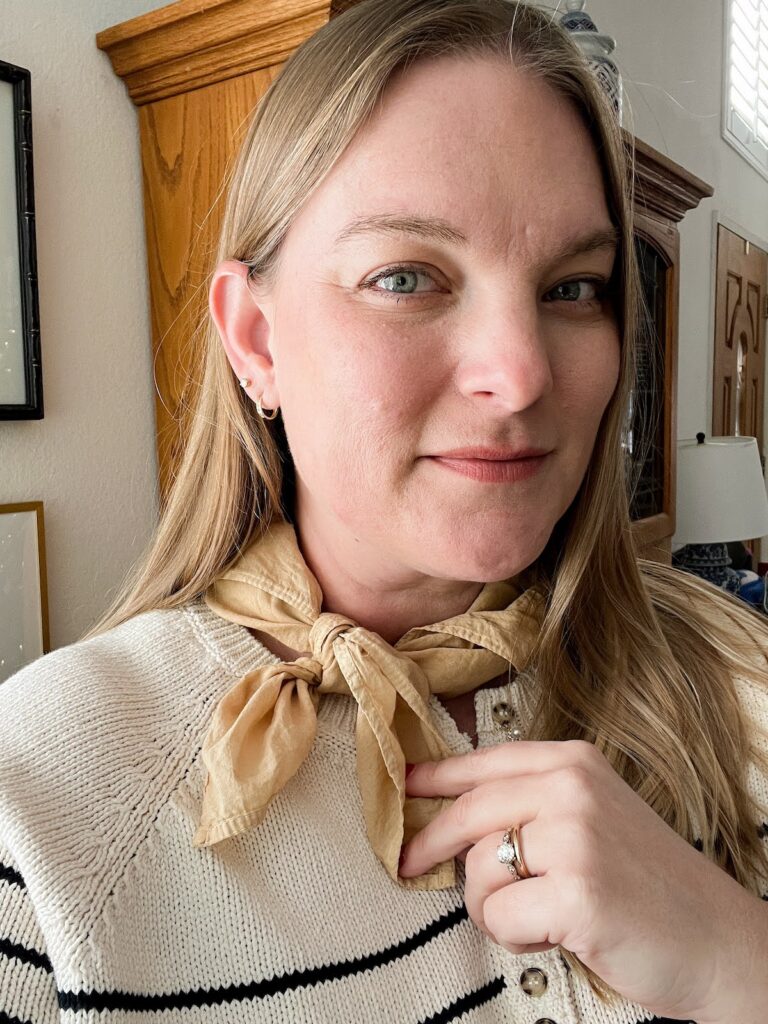 A blonde white woman in her late 30s (me!) smiles softly at the camera. She's wearing a striped navy and cream sweater and a tan neckerchief.