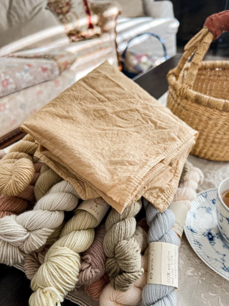 A folded tan neckerchief sits on top of a pile of pastel mini skeins of yarn. A grass basket sits empty in the background.