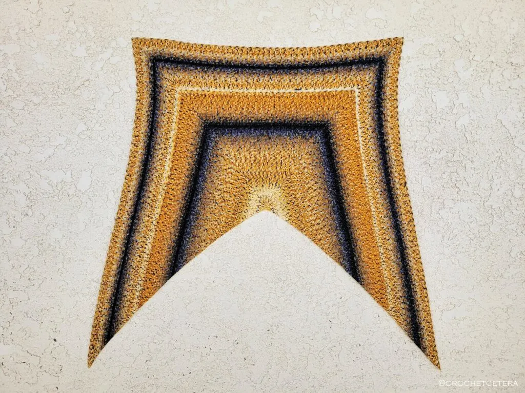 A concentric square shawl with gold, cream, and black stripes is laid flat on a stucco background.