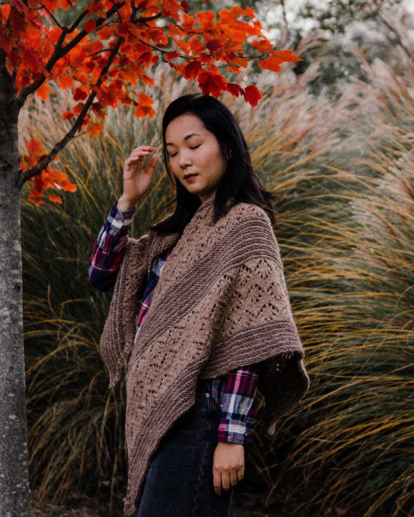 An Asian-American person stands beneath a tree full of rich red foliage, draped in a thick, warm shawl with alternating bands of lace and garter stitch.