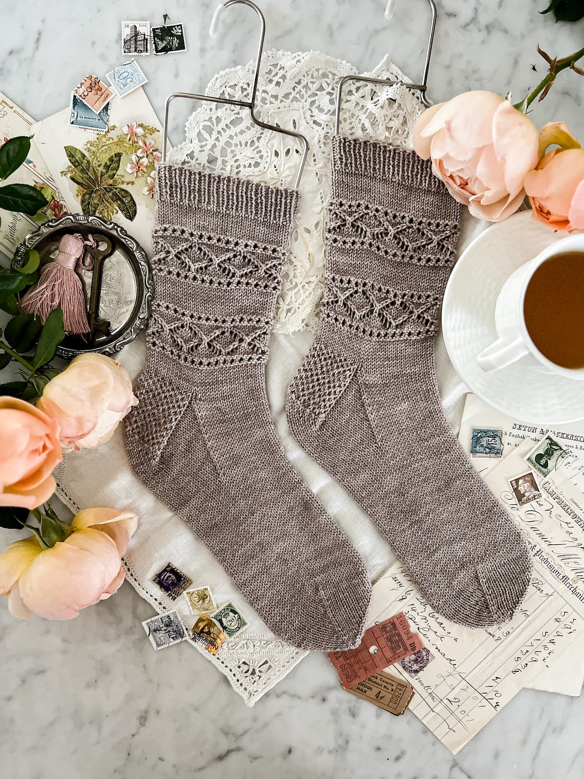 A flatlay photograph of two gray, handknit socks stretched on wire sock blockers. They have lacy bands around the legs and the toes are both pointing to the right. They're surrounded by peach roses and antique paper ephemera.
