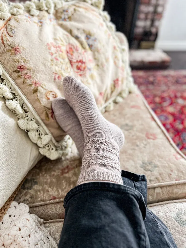 A pair of feet wearing gray, handknit socks are stretched out on an antique sofa and crossed at the ankles.