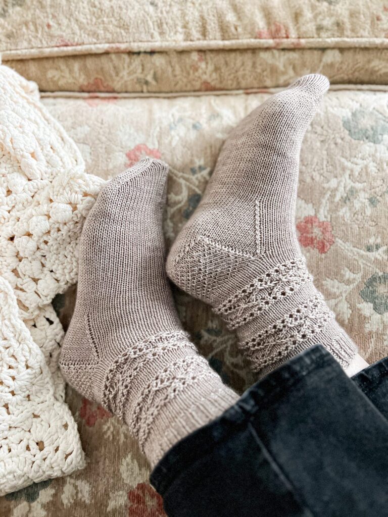 A pair of feet wearing gray, handknit socks are photographed from the side on an antique sofa. The socks have lace bands around the ankles.