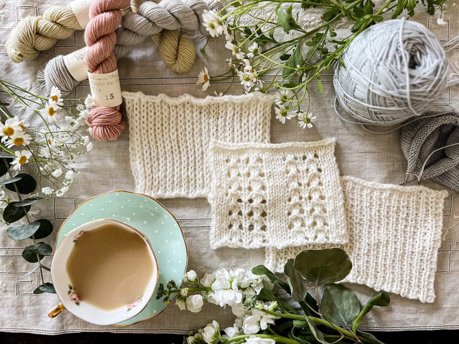 Three swatches of white knit fabric showing different slipped stitches are laid out in a diagonal on a cream linen background. They're surrounded by pastel yarn, fresh flowers, and a teacup full of milky tea.
