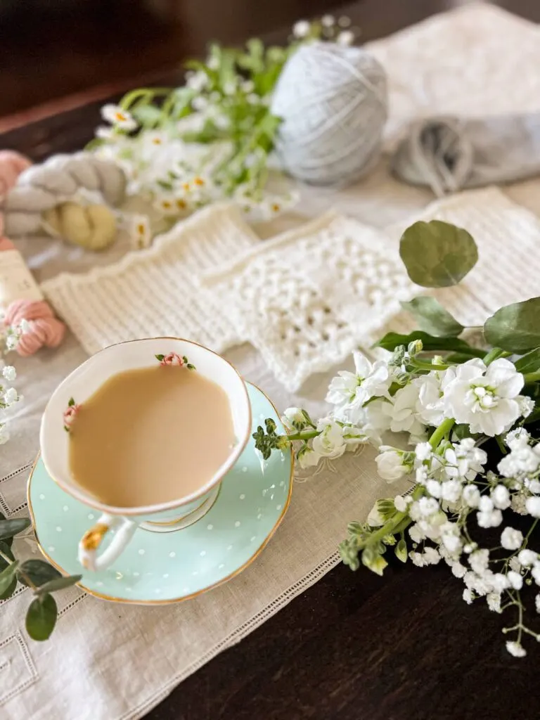 A teacup full of milky tea fills the foreground in the lower right corner of the photo. Blurred in the background are three swatches of white knit fabric, some fresh flowers, and pastel yarn.