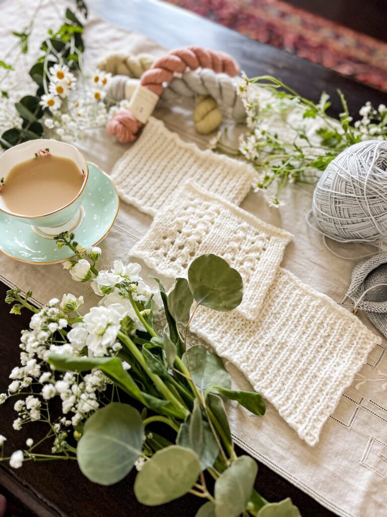 A high-angle photo of three white swatches of knit fabric laid out on a diagonal and photographed from the lower right side of the diagonal. There are fresh flowers, pastel yarn, and a teacup full of milky tea.