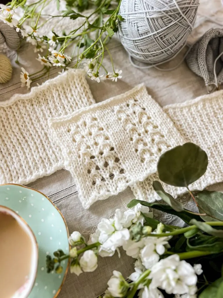 A swatch of knit fabric with stitches slipped in different styles is laid on top of two other swatches of white knit fabric. They're arranged diagonally and surrounded by fresh flowers and pastel yarn.