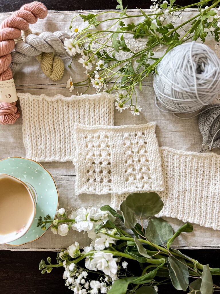 A photo showing three swatches of white knitting with different slipped stitch textures. They're surrounded by fresh flowers, mini skeins of pastel yarn, and a teacup full of milky tea.