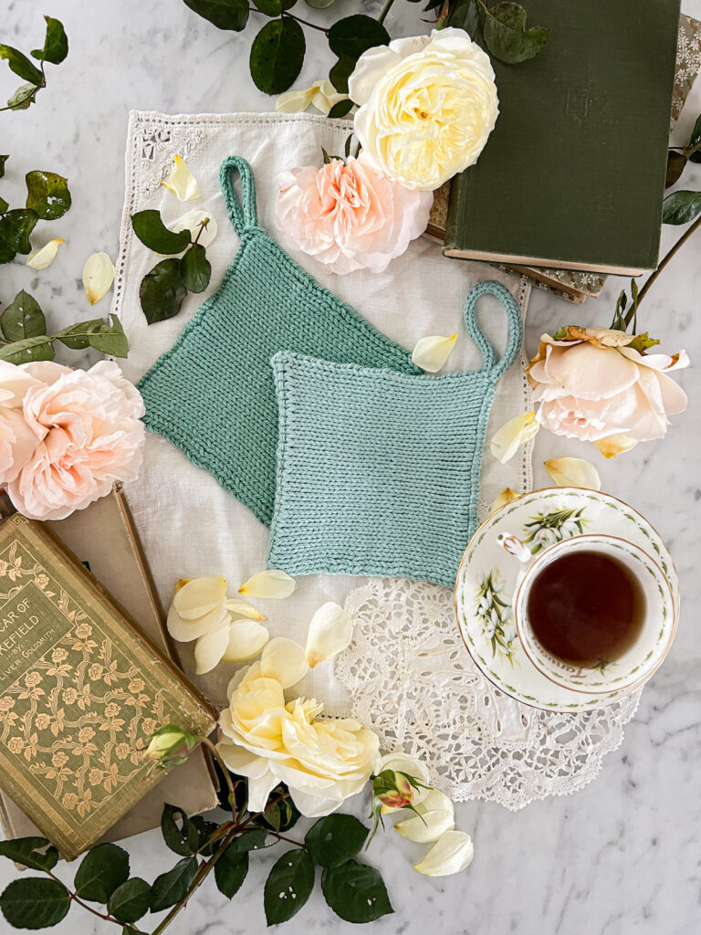 A flatlay photograph showing two handknit potholders in teal and seafoam green. They're surrounded by pink and yellow roses, a vintage teacup full of tea, and vintage books.