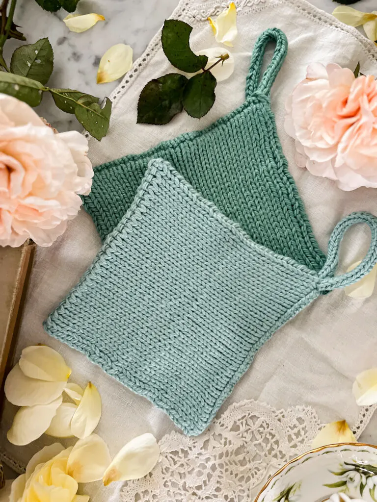 A top-down, zoomed-in photo of a seafoam green, handknit potholder sitting on top of a darker teal potholder knit in the same style. They're surrounded by pink and yellow roses.