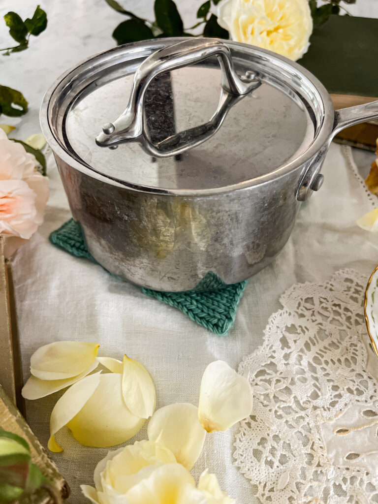 A steel sauce pan with matching lid (speckled from heavy use and hard water) sits on top of a teal handknit pot holder. They're surrounded by yellow and pink roses.