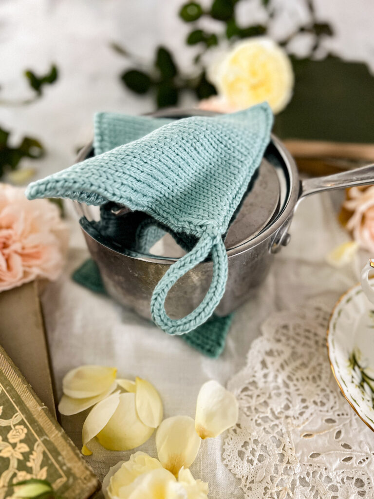 A seafoam green knit potholder is curved over the lid of a steel sauce pan. The pan sits on top of another knit potholder that is serving as a trivet. They're surrounded by pink and yellow roses and vintage books.