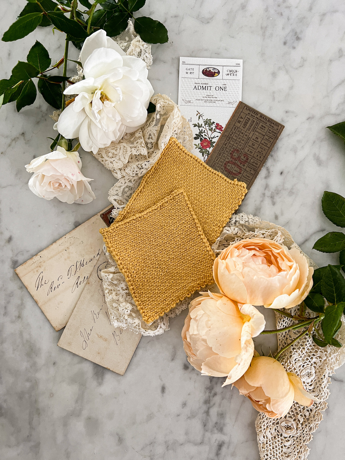 A flatlay photograph showing two yellow knit swatches photographed from the top down. Both are knit in linen stitch. The top swatch was knit with larger needles and therefore has a looser gauge. They're surrounded by white and peach roses, antique lace, and paper ephemera.