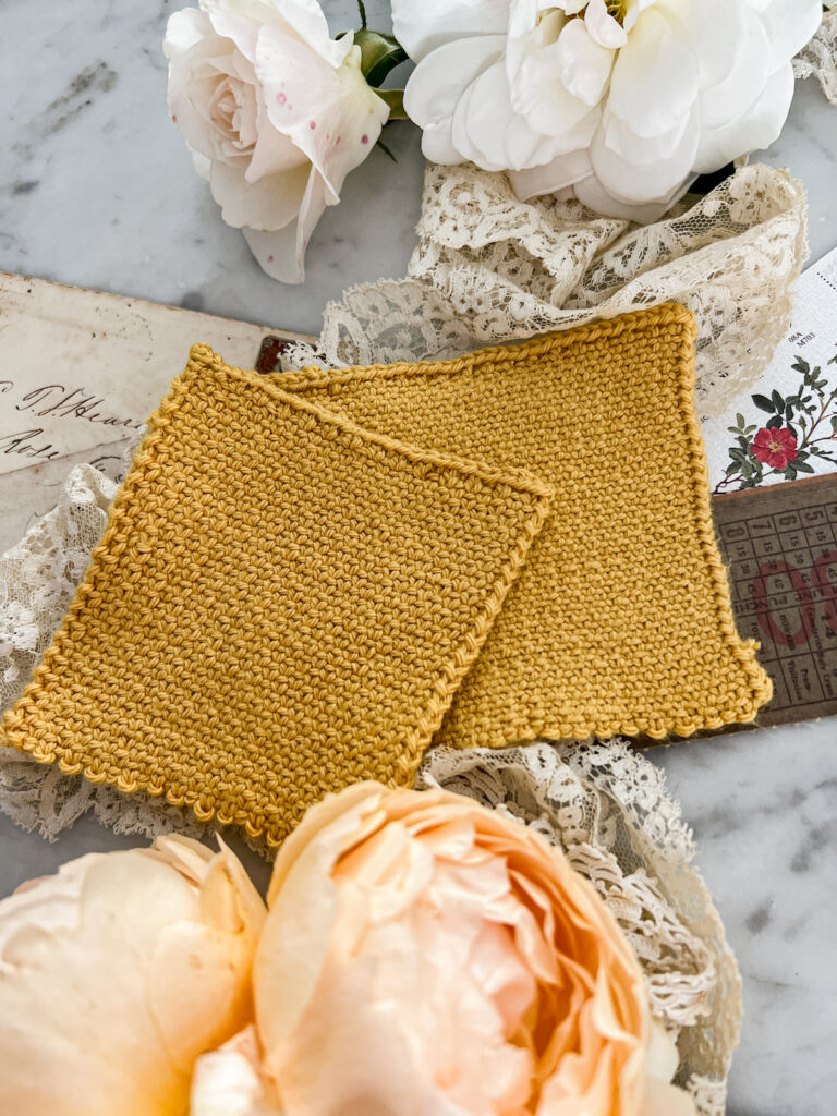 Another angle showing two linen stitch swatches laid out on top of each other. The top swatch was knit with larger needles. They're surrounded by white and peach roses, antique lace, and paper ephemera.