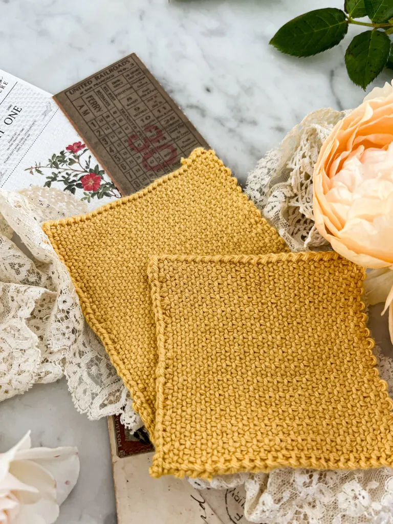Two yellow knit swatches in linen stitch are laid out in an overlapping manner, with one peeking out from behind the other. They're surrounded by white and peach roses, antique lace, and paper ephemera.